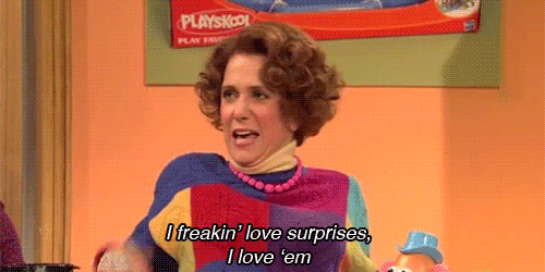 SNL gif. Kirsten Wiig is dressed like a woman in the 70s, with curly short hair, turtleneck, thick pink pearls, and patchwork sweater that are all different colors. She claps her hands eagerly and leans forward while saying, "I freakin' love surprise. I love em."