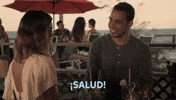 TV gif. Victor Rasuk as Daniel and Nathalie Kelley as Noa in The Baker and the Beauty tip their drinks to each other as he smiles at her and they say, "Salud! Salud! Salud" back and forth to each other.