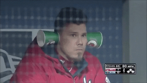 Sports gif. Jose Lobaton with the Washington Nationals Baseball team is chewing sunflower seeds and has two cups stuck on his ears to amplify the noise. He spots the camera and he holds two more cups to his eyes, looking at us.