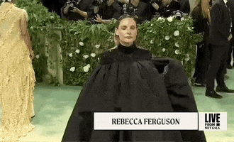 Met Gala 2024 gif. Rebecca Ferguson tossing her Thom Browne black silk moiré cape to one side, revealing embroidered black ravens against a sky-blue inner lining, inspired by Edgar Allan Poe. Her black grown features sequined shoulders and green and blue Swarovski crystals embroidered into flowers. 