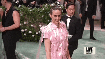Met Gala 2024 gif. Camera zooms out on Sarah Paulson's pale pink short-sleeved Prada gown with a straight silhouette and long, thin panels of tulle that extend from her shoulders to the ground. The dress is covered in columns of pale pink satin bows that catch the light.