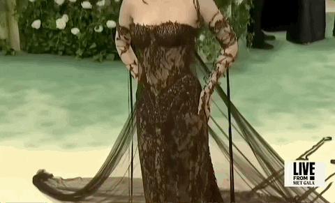 Met Gala 2024 gif. Closeup of the top of Emma Chamberlain's gothic-style Jean Paul Gaultier sleeveless black lace gown that features thin dark brown strands of lace weaved around her arms resembling brambles and ferns. She's wearing dark brown eye makeup and lipstick and a diamond collar necklace. 