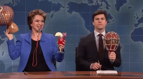SNL gif. Holding a cocktail, shaking a beaded maraca, and blowing a coach's whistle, Maya Rudolph sits next to Colin Jost on Weekend Update, who shyly shakes a maraca.