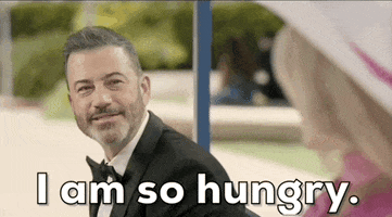 Oscars 2024 GIF. Jimmy Kimmel sits next to Margot Robbie as Barbie on the bus stop bench and he shakes his head seriously and solemnly as he says, "I am so hungry" as he parodies the bus stop bench scene.