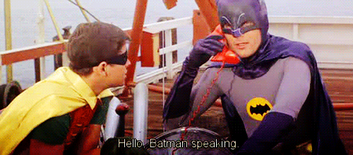 TV gif. Adam West as Batman and Burt Ward as Robin sit on a boat. Batman holds a red phone up to his ear and says, “Hello, Batman speaking.” 