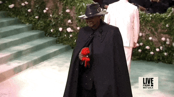 Met Gala 2024 gif. Usher, wearing an all-black custom Alexander McQueen ensemble, holds and lifts a red paper flower.