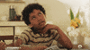 TV gf. Adrian Groulx as young Dwayne Johnson on Young Rock sits at a kitchen table. He has his hand on his cheek and he flops to his side, leaning onto the table, as he rolls his eyes and sighs.