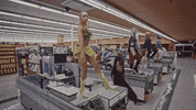 Music video gif. A scene from Tinashe's music video for "Needs" shows Tinashe and six of her dancers loosely kicking their legs and swinging their arms with swagger on the top of checkout counters in an empty grocery store. 