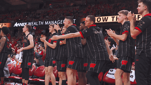 Sports gif. Members of the Wisconsin Badgers basketball team clap and dance on the sidelines as they watch the game. One guy in the center does an awkward little dance, tapping his feet with bent knees as he tosses his straight arms back. 