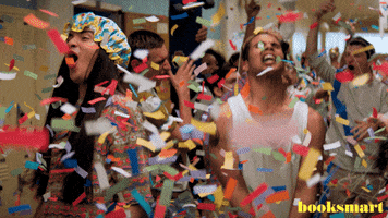 Movie gif. Eduardo Franco as Theo in Booksmart, wearing a shower bonnet, joins his classmates in celebrating in the hallway as confetti streams down from the ceiling.
