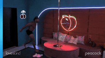 Reality TV gif. Jesse from Love Island, dressed only in a bandanna and boxer briefs, jumps onto and swings around a pole in a room decorated with neon designs and club lighting.
