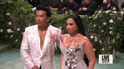 Met Gala 2024 gif. Pabral Gurung poses with Demi Lovato, wearing his smokey lavender gown embellished with silver flowers, as he adjusts and waves.