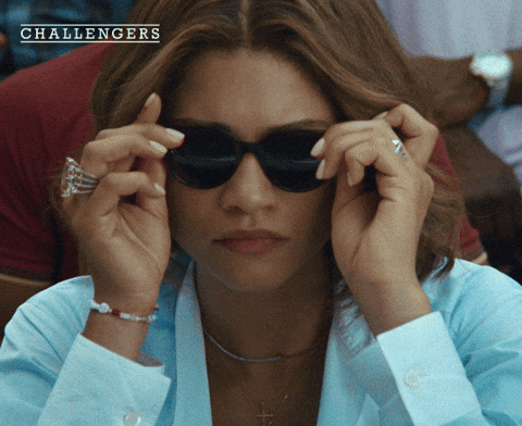 Movie gif. A shot from the movie "Challengers." Tashi Donaldson puts on a pair of large sunglasses, then sits back in her seat. She frowns slightly. 