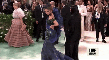 Met Gala 2024 gif. Zendaya glides across the red carpet, turning her head for a dark stare towards camera. She's wearing a midnight blue and dark teal dress with a large chiffon puffed sleeve over her right shoulder and net mesh and tulle feathered fascinator. The dress is trumpet style with a dramatic flare at the base.