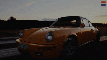 Video gif. Ruf CTR car rides smoothly along an empty road as the sun sets cinematically in the background, casting the car in a soft orange glow.