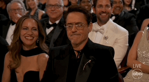 Oscars 2024 GIF. Robert Downey Junior, seated at the Oscars, cranks the gesture for camera casually but emphatically.