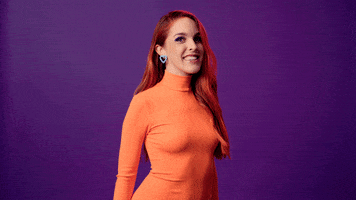 Celebrity gif. Amarna Miller stands in front of a solid purple background wearing an orange turtleneck. She points upward with a playfully excited smile.
