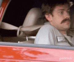 The Office gif. John Krasinski as Jim sits in the passenger seat of a parked car, wearing a fake moustache and gray jumpsuit. He looks at us with discomfort as he reclines the chair out of sight behind the car door.