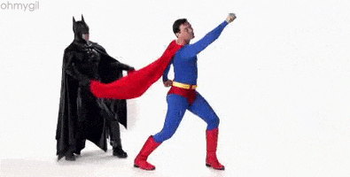 Video gif. 2 live action superheroes superimposed on a white background. Superman stands in a flying pose while Batman stands behind him, flapping his cape as if it were flowing in the wind. 
