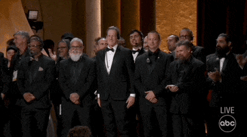 Oscars 2024 GIF. A large group of Teamsters, Gaffers, and Grips come onstage in suits and grin at the crowd as the audience cheers for them. 