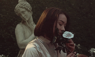 Music video gif. Mitski deeply smells a flower then exhales contently in a statue garden in the music video, "Stay Soft."