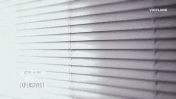 Video gif. A man peeks through a set of window blinds and leans in to get a better look as if he was trying to spy on someone or something. 