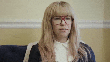 TV gif. Carrie Brownstein on Portlandia wears a blonde wig and big aviator glasses. She looks around and shrugs her shoulders.