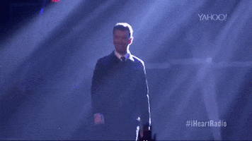 Celebrity gif. Walking on an ethereally lit stage at an I Heart Radio festival, Sam Smith smiles and waves at the audience.