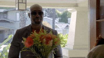 TV gif. Morris Chestnut as Beaumont in Rosewood wears sunglasses and holds a bouquet of flowers as he tips his head in a flirty nod.