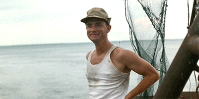 Movie gif. Tom Hanks as Forrest Gump stands on a boat with a wide smile on his face. His eyes beam as he gazes ahead, raising a hand and waving energetically. 