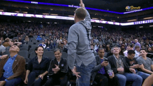 Sports gif. Jason Williams, former Sacramento Kings guard, stands up and waves as people in the audience clap for him.