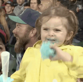 Video gif. Toddler girl clasps a fistful of blue cotton candy that's also smeared around her mouth. She smiles maniacally through gritted teeth, her eyes growing big, as she stands on a woman's lap in a stadium full of people.