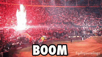 Sports gif. At the edge of dirt bike arena, near a crowded audience, a fiery explosion bursts upward as a motocross driver exits a ramp and soars high above the arena.