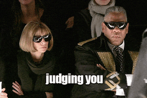 Photo gif. Anna Wintour sits in the audience of a runway show with her arms crossed and sunglasses on that makes her expression more serious. Andres Leon Talley looks at us with a similar expression and crossed arms. The text, “Judging you,” flashes bright pink and white.