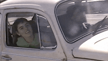Music Video gif. Hazel English in the Never Going Home music video leans her head out of the window of an old Volkswagen Beetle as a man drives her down a highway.