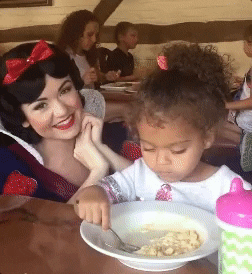 Video gif. A little girl sits at a table with her bowl and sippy cup. She stares blankly straight ahead, as a woman dressed as Snow White tries to say hi to her, smiling at her and us. The girl continues to ignore her.