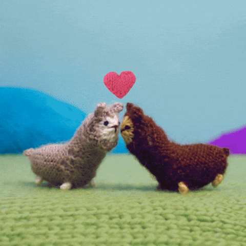 Stop-motion gif. Knitted scene with a gray alpaca and a brown alpaca who turn to face each other and kiss, as pink knitted hearts fly up between them.