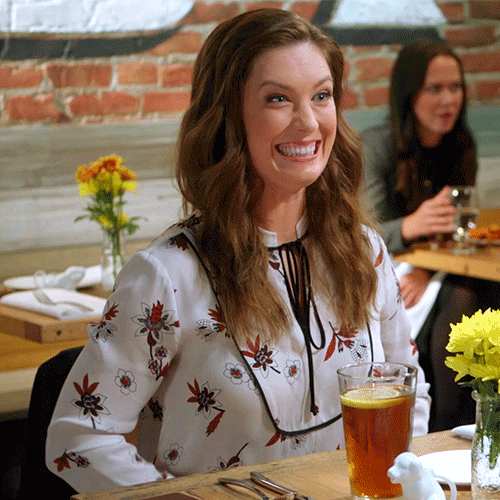 TV gif. Briga Heelan as Katie in Great News sits across from someone at a dining table and beams as she claps excitedly.