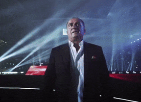 Sports gif. Faith Terim, former Galatasaray coach, walks heroically in a tuxedo through a dark stadium lit up from the crowd and LED stage lights. Cameras flash as he puts his hand over his heart and acknowledges the crowd. 