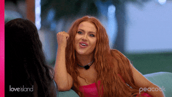 Reality TV gif. Sydney of Love Island leans onto the arm of a couch, raising her eyebrows and expressing surprise while listening to someone.