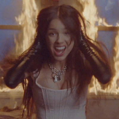 Music Video gif. Olivia Rodrigo in the Good Four U music video stands in front of a burning window as she screams at us. Her black leather gloved hands run down her face like she’s emulating the famous scream painting.