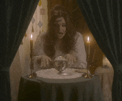 Video gif. Woman dressed as a fortune teller sits at a circular table with candles and tarot cards. She waves a hand over a crystal ball and text appears, "I seeee... That you're going to come into some money. A lot of money." She looks up and gives us an cheesy, exaggerated grin.