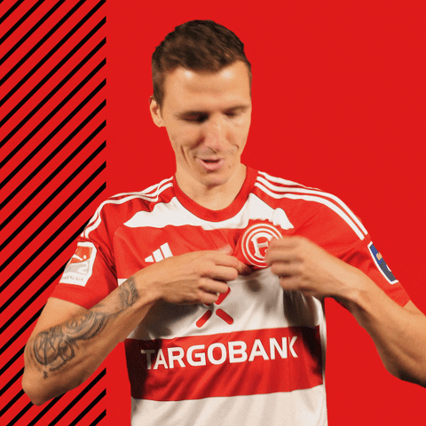 Video gif. Marcel Sobottka of Fortuna Dusseldorf lifts his shirt to gnaw in a silly animated way at the Fortuna logo on his chest. 