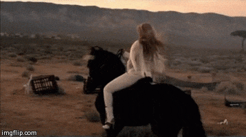Video gif. In slow motion, a woman wearing white pants and a fringed white jacket rides a dark horse across a desert landscape.