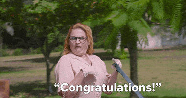 Video gif. Woman wearing cat-eye glasses walking a dog smiles with pride as she holds up a gloved hand and says, “Congratulations.”